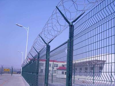 Green color curvy welded mesh fences with razor wires are installed outside of the prison.