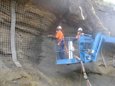 Two laborers are spraying concrete on a concavity of a mountain.